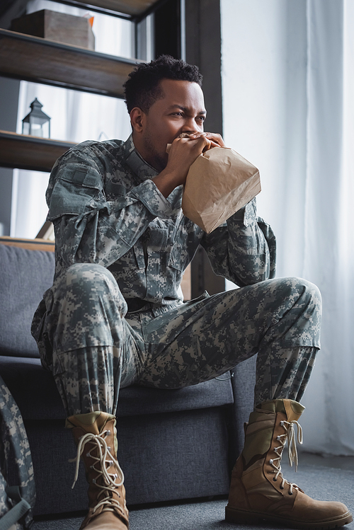 african american soldier in military uniform breathing with paper bag while having panic attack at home
