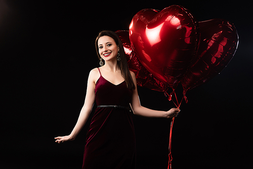 smiling woman in dress holding heart-shaped balloons in 14 february isolated on black