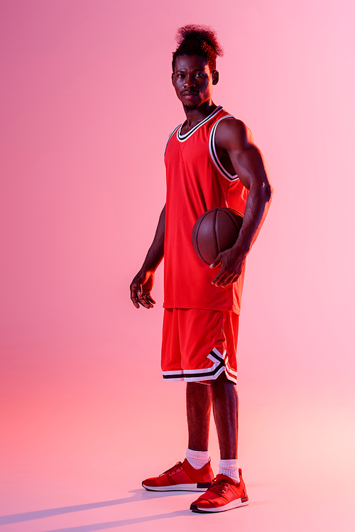 serious african american basketball player  while holding ball on pink background with lighting and gradient