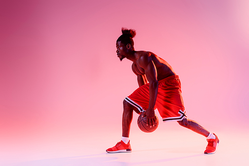 shirtless, muscular african american sportsman playing basketball on pink and purple gradient background with lighting