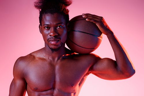 confident, shirtless african american basketball player holding ball and  on pink background with gradient