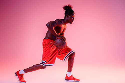 african american sportsman in red short playing basketball on pink and purple gradient background