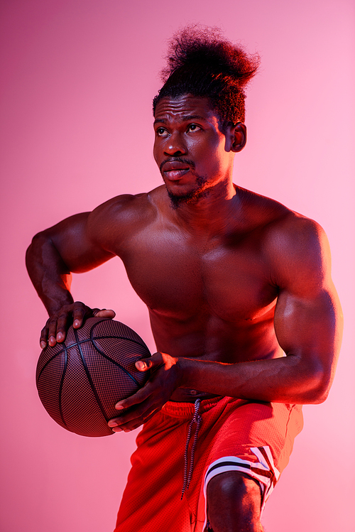 serious african american basketball player holding ball and looking away on pink and purple gradient background