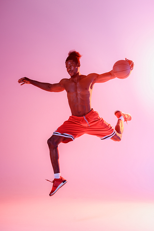shirtless african american sportsman in red shorts jumping while playing basketball on pink and purple gradient background