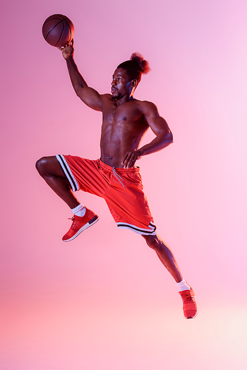 athletic african american sportsman in red shorts playing basketball on pink and purple gradient background