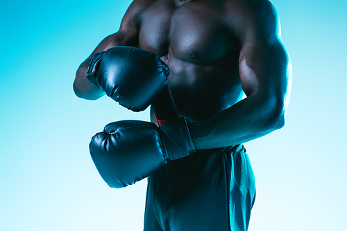 cropped view of shirtless, muscular african american sportsman in boxing gloves on blue background