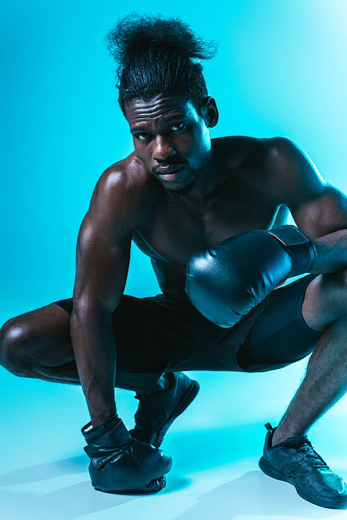 confident, muscular african american sportsman in boxing gloves posing at camera on blue background