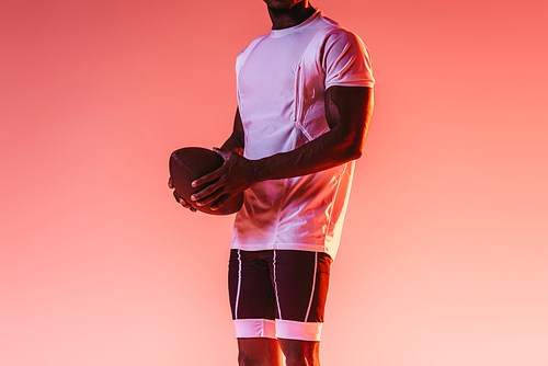 partial view of african american sportsman holding rugby ball on pink background with gradient and lighting