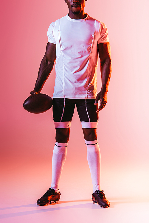 cropped view of african american sportsman holding rugby ball on pink background with gradient and lighting