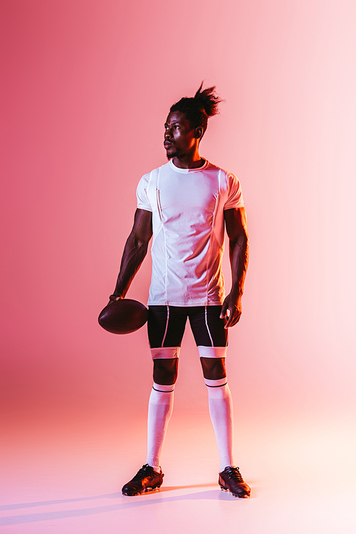 handsome african american sportsman looking away while holding rugby ball on pink background with gradient and lighting
