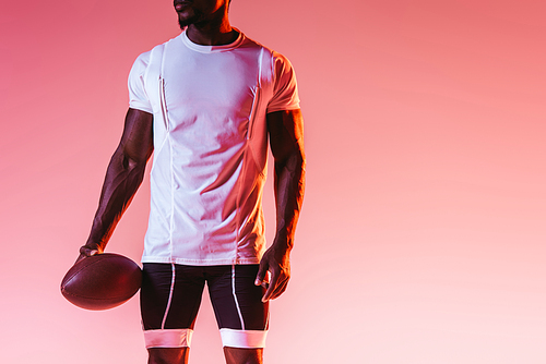 cropped view of african american sportsman holding rugby ball on pink background with gradient and lighting