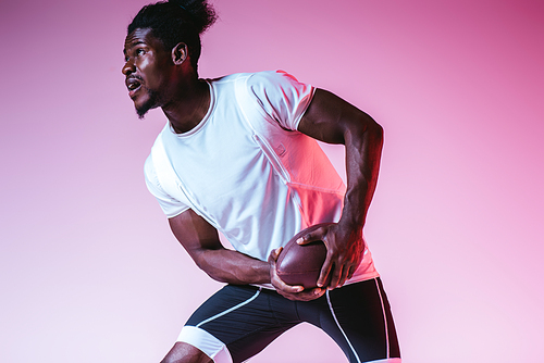 excited african american sportsman playing american football on purple background with gradient