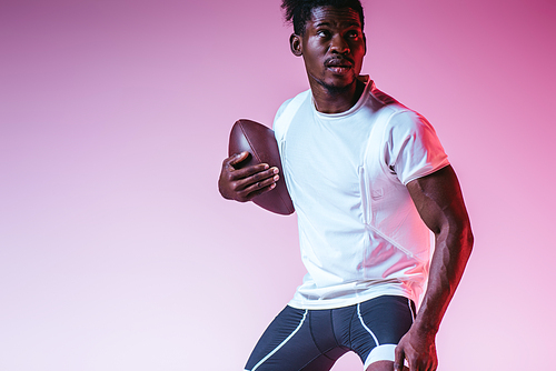 handsome african american sportsman playing american football on purple background with gradient
