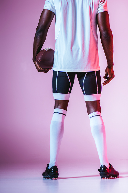 back view of african american sportsman holding rugby ball on purple background with gradient and lighting