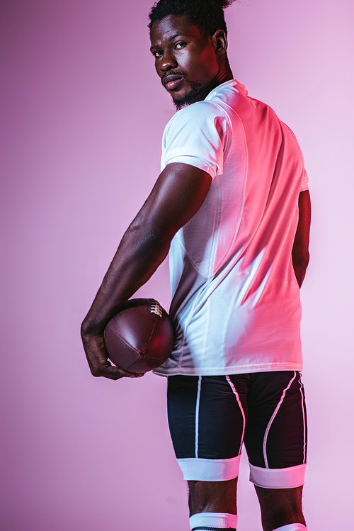 confident african american sportsman  while holding rugby ball on purple background with gradient