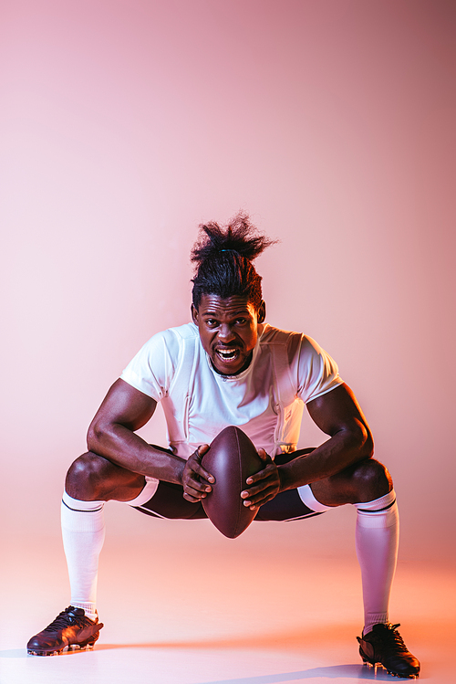 excited african american sportsman yelling while playing american football on pink background with gradient and lighting