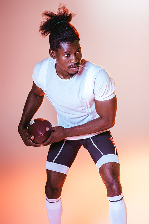 confident african american sportsman playing american football on pink background with gradient and yellow lighting