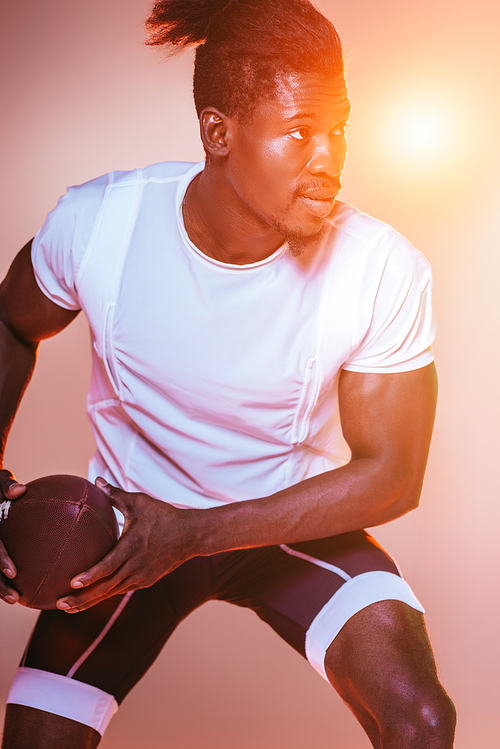 handsome african american sportsman playing american football on pink background with gradient and yellow lighting