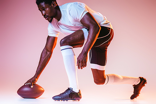young african american sportsman playing american football on pink background with gradient and yellow lighting