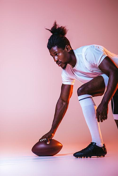 young african american sportsman playing american football on pink background with gradient and lighting
