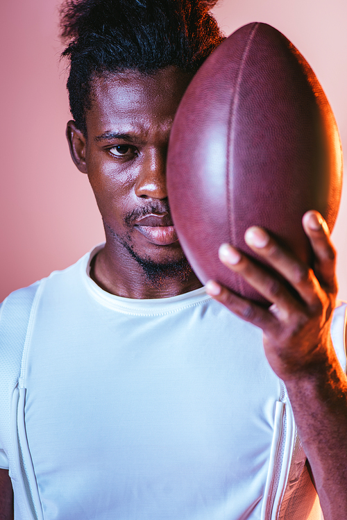 serious african american sportsman  while holding rugby ball on pink background with lighting