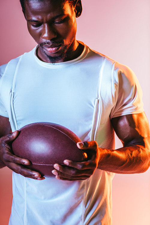 handsome, young african american sportsman holding rugby ball on pink background with lighting