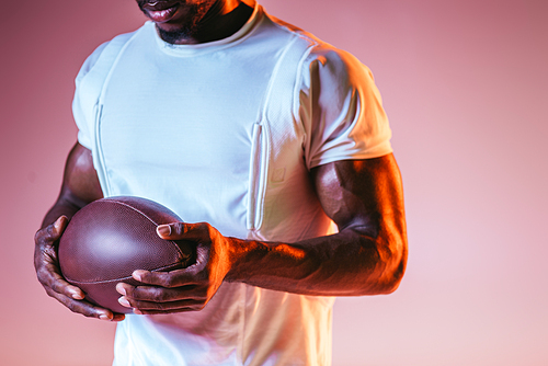 cropped view of afican american sportsman holding rugby ball on pink background with gradient and lighting