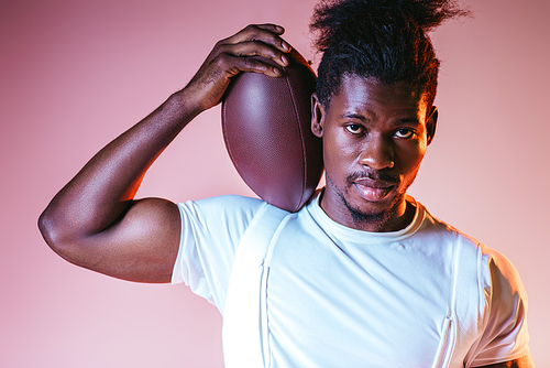 handsome african american sportsman  while holding rugby ball on pink background with gradient