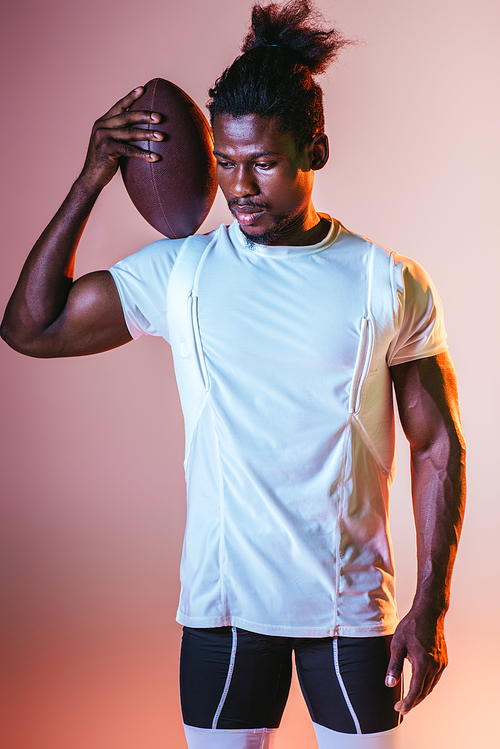young african american sportsman holding rugby ball on pink background with gradient and lighting