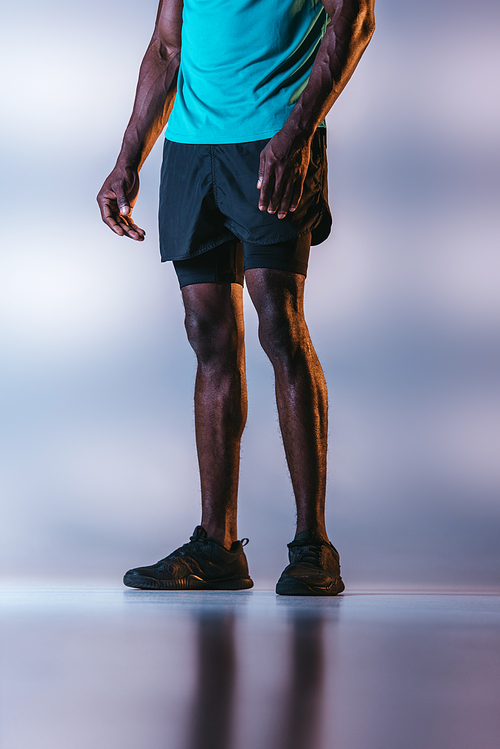 cropped view of african american sportsman in shorts and sneakers on grey background with lighting