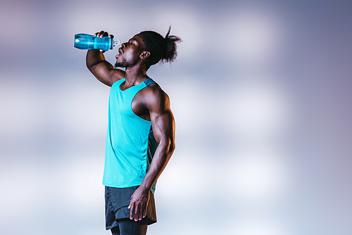 young african american sportsman drinking from sports bottle on grey background with lighting