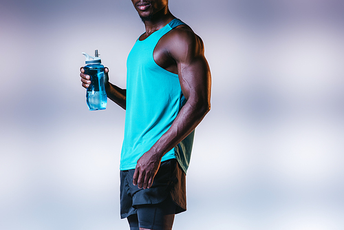 cropped view of muscular african american sportsman holding sports bottle on grey background with lighting