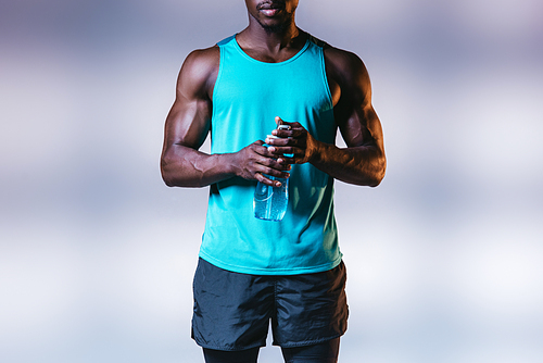 partial view of muscular african american sportsman holding sports bottle on grey background with lighting