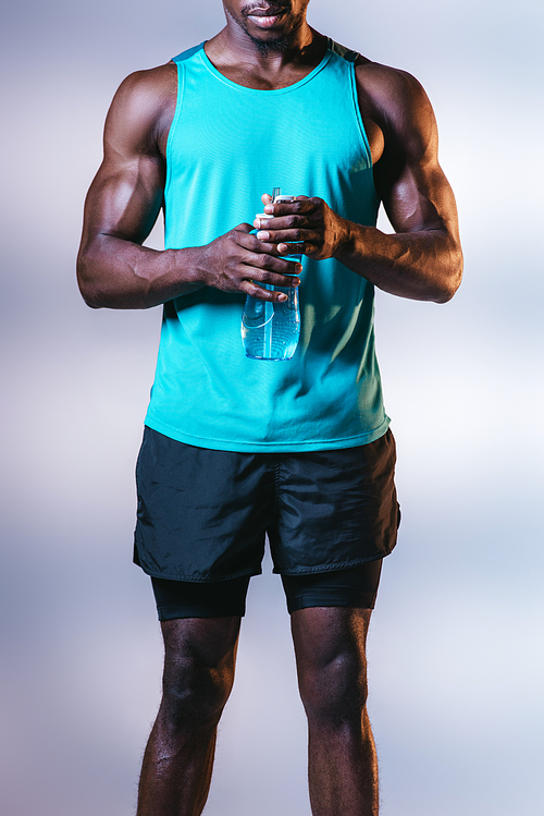 partial view of african american sportsman in shorts holding sports bottle on grey background with lighting