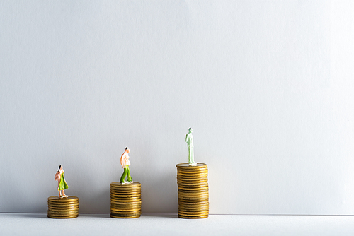 Three people figures on stacked golden coins on white surface on grey background, concept of financial equality