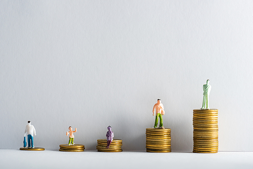 Plastic people figures on stacked golden coins on white surface on grey background, concept of financial equality