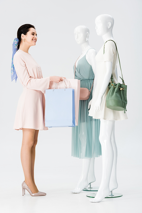 smiling young woman with shopping bags near mannequins isolated on grey