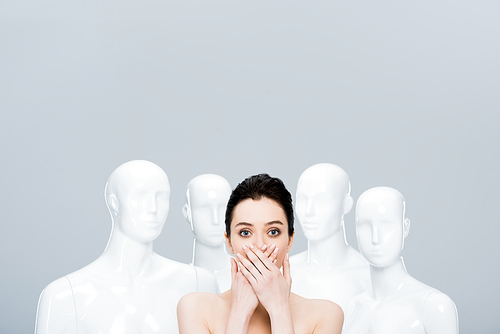 beautiful young woman covering mouth with hands while posing near mannequins isolated on grey