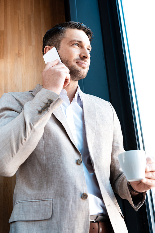 smiling businessman holding cup of coffee and talking on smartphone in cafe