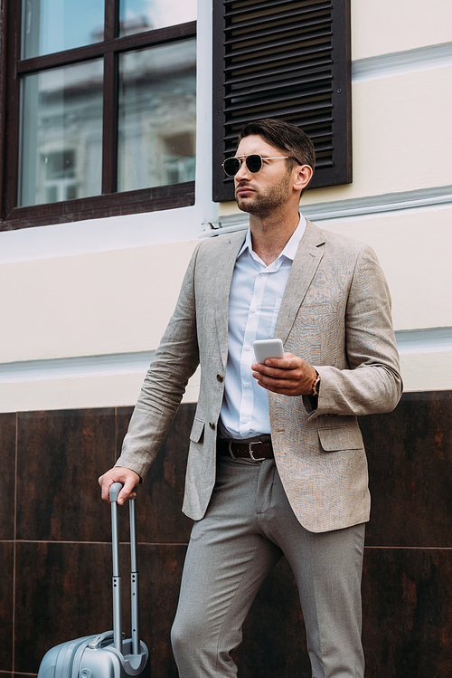 businessman in sunglasses holding suitcase and smartphone on street