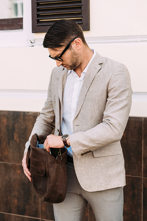 serious businessman in wristwatch holding leather bag on street