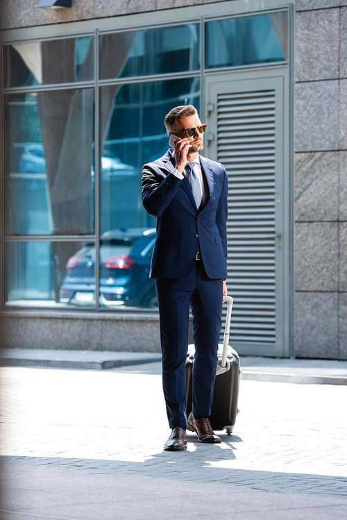 handsome man in suit and glasses holding suitcase and talking on smartphone