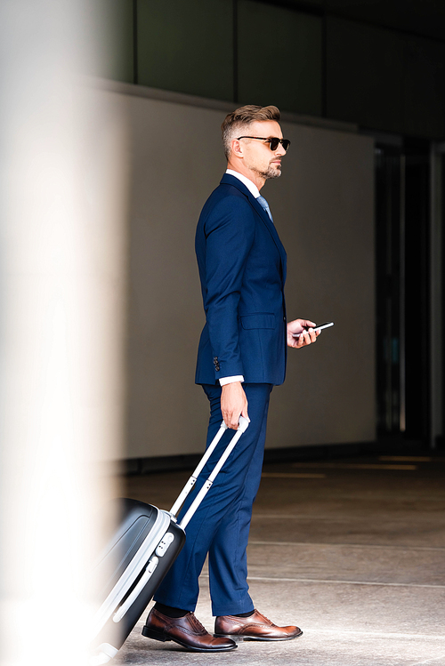 handsome businessman in suit and glasses holding suitcase and smartphone