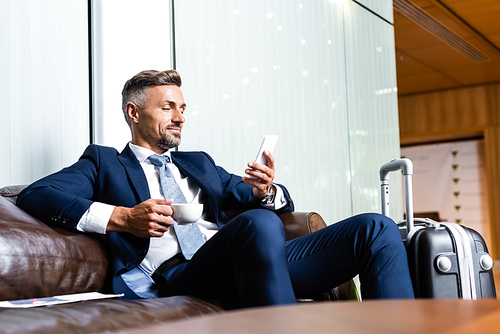 handsome businessman in suit using smartphone and holding cup