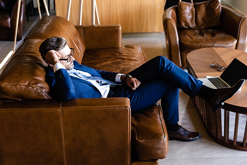 side view of handsome businessman in suit and glasses sitting on sofa