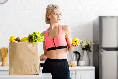 attractive blonde woman in sportswear holding apple near paper bag with groceries