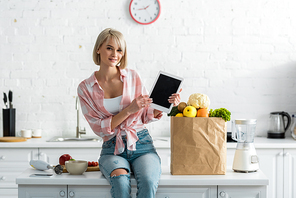 beautiful blonde woman holding digital tablet with blank screen near paper bag with ingredients