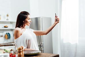 Charming young woman taking selfie near table in kitchen