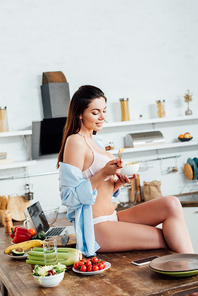 Sexy girl in underwear and shirt sitting on table and eating fruit salad