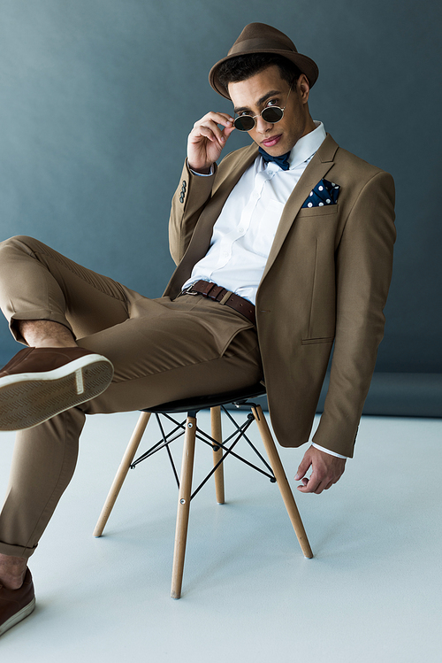 stylish mixed race man in suit and sunglasses sitting on chair and  on grey and white